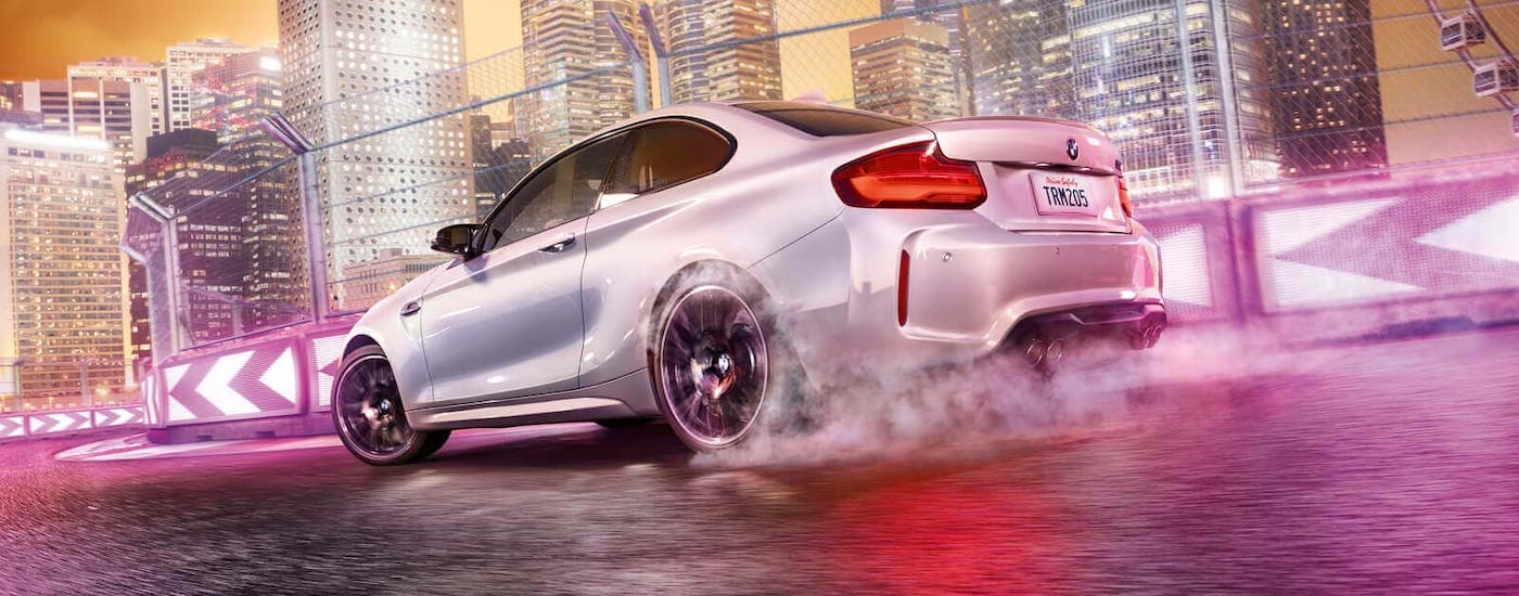 A silver 2021 BMW M2 Competition Coupe is driving on a racetrack in front of a vibrant pink and yellow city.