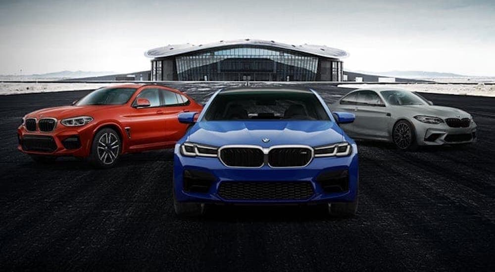 A red, a blue, and a silver BMW M Series sedans and coupes are parked in front of a hangar.
