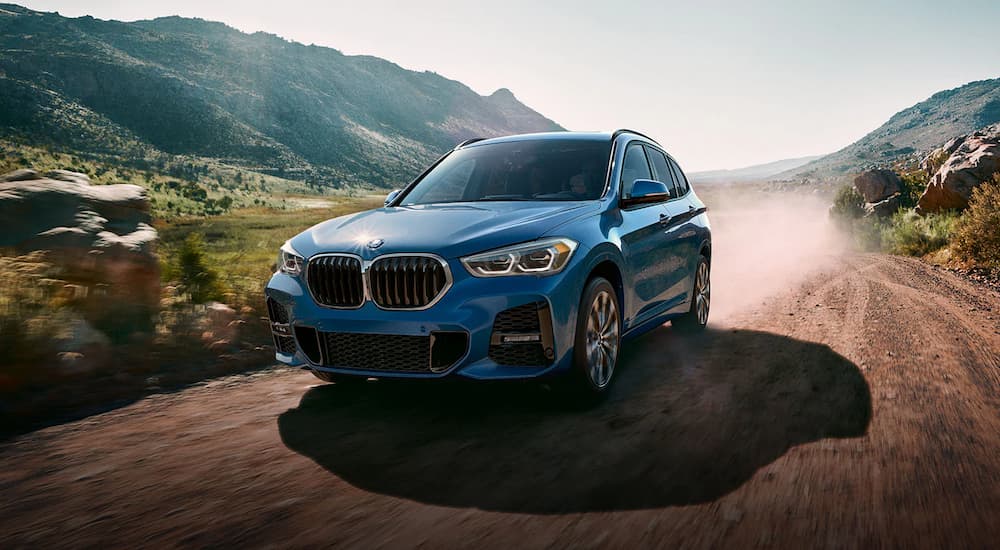A blue 2022 BMW X1 M Sport is shown from a front angle driving down a dirt road after researching BMW lease deals.